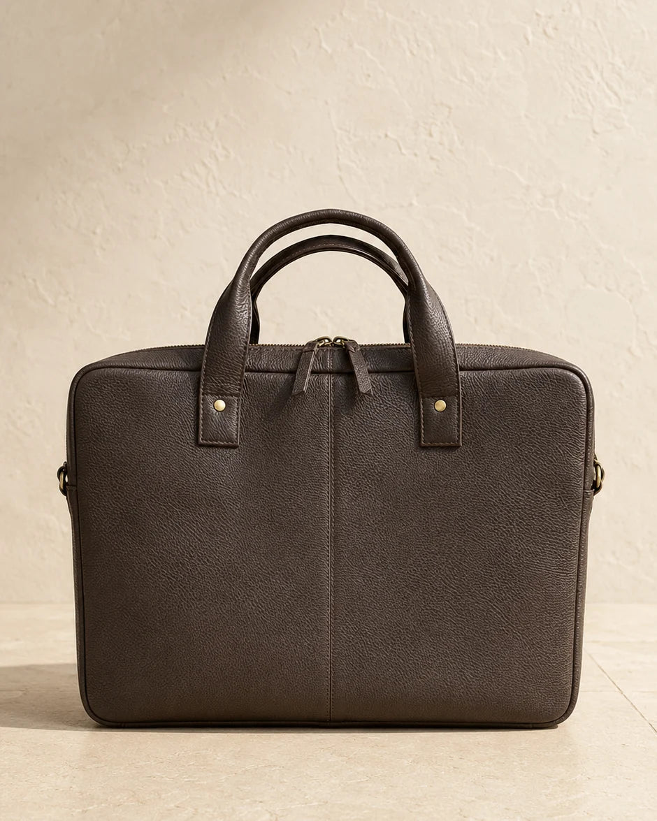 A dark brown leather briefcase with dual top handles and gold-tone hardware, photographed against a textured beige wall and floor.