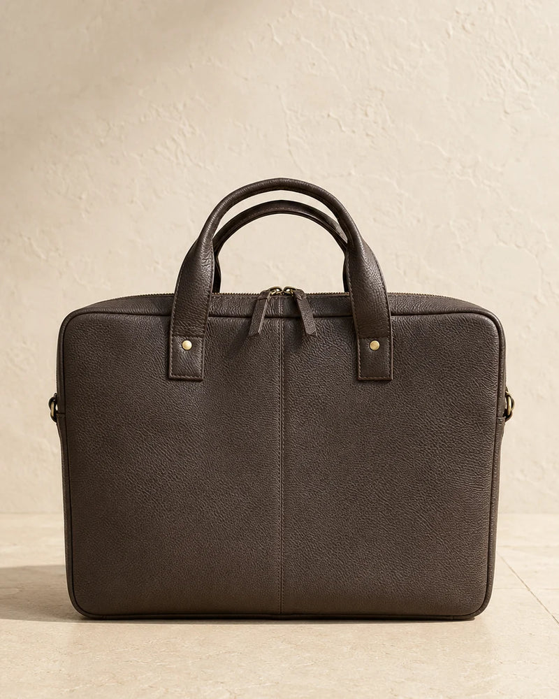 A dark brown leather briefcase with dual top handles and gold-tone hardware, photographed against a textured beige wall and floor.